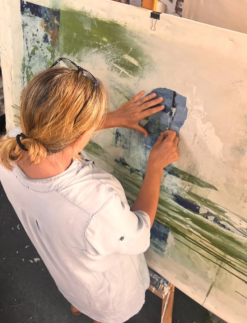 Aline Chevalier à l’atelier, les mains posant un fragment de papier bleu sur une œuvre en cours.